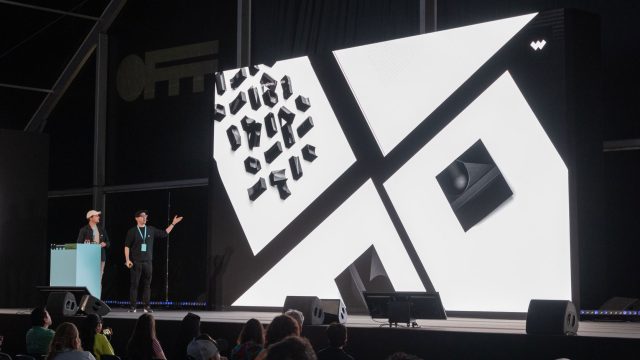 Two people stand on a stage next to a large screen showing abstract black and white geometric shapes during the CD at JVG, as the audience watches the OFFF x Stash Interview with Javier Vallejo. Frame from Stash Magazine article.