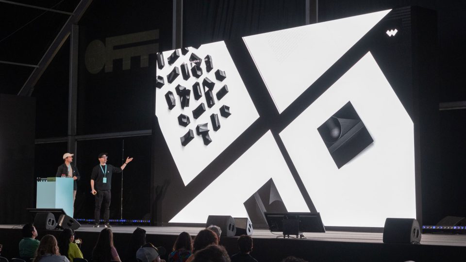 Two people stand on a stage next to a large screen showing abstract black and white geometric shapes during the CD at JVG, as the audience watches the OFFF x Stash Interview with Javier Vallejo. Frame from Stash Magazine article.