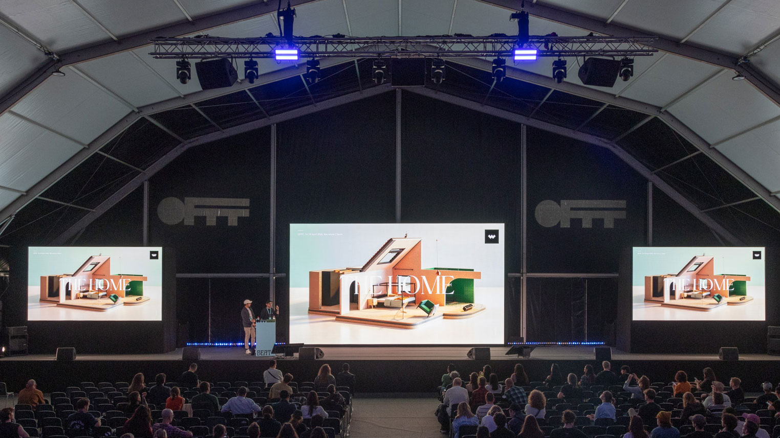 A large indoor stage at OFFF features three screens displaying the word WELCOME and two people presenting, while attendees sit spaced out in rows under a high, tent-like ceiling—Javier Vallejo's name appears among the visuals curated by Stash. Frame from Stash Magazine article.