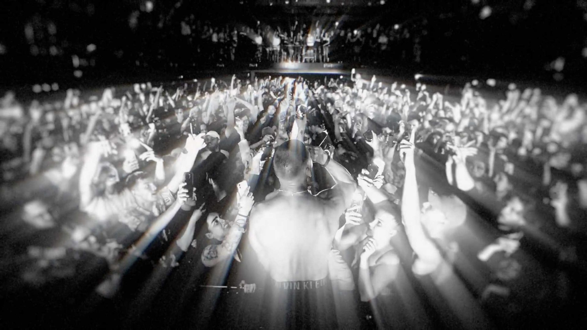 A black and white image of a crowded concert, with people raising their hands and phones. Light beams radiate from the center, highlighting the audience and creating a dramatic, immersive effect—true to the energy of Never Sit Still. Frame from Stash Magazine article.