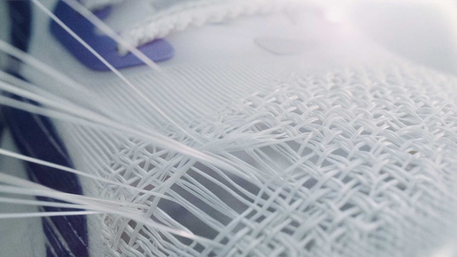 Close-up of a white Olympikus Pace running shoe, focusing on the woven mesh texture and synthetic fibers, with some threads pulled and frayed, and hints of purple accents in the background, as seen in the product film by Miagui. Frame from Stash Magazine article.
