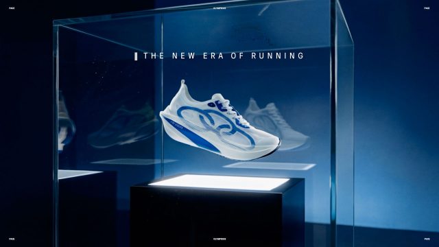 A futuristic white and blue Olympikus Pace running shoe is displayed in a glass case under spotlight, with the text THE NEW ERA OF RUNNING above it on a dark blue background, inspired by the Olympikus Pace running shoe product film by Miagui. Frame from Stash Magazine article.