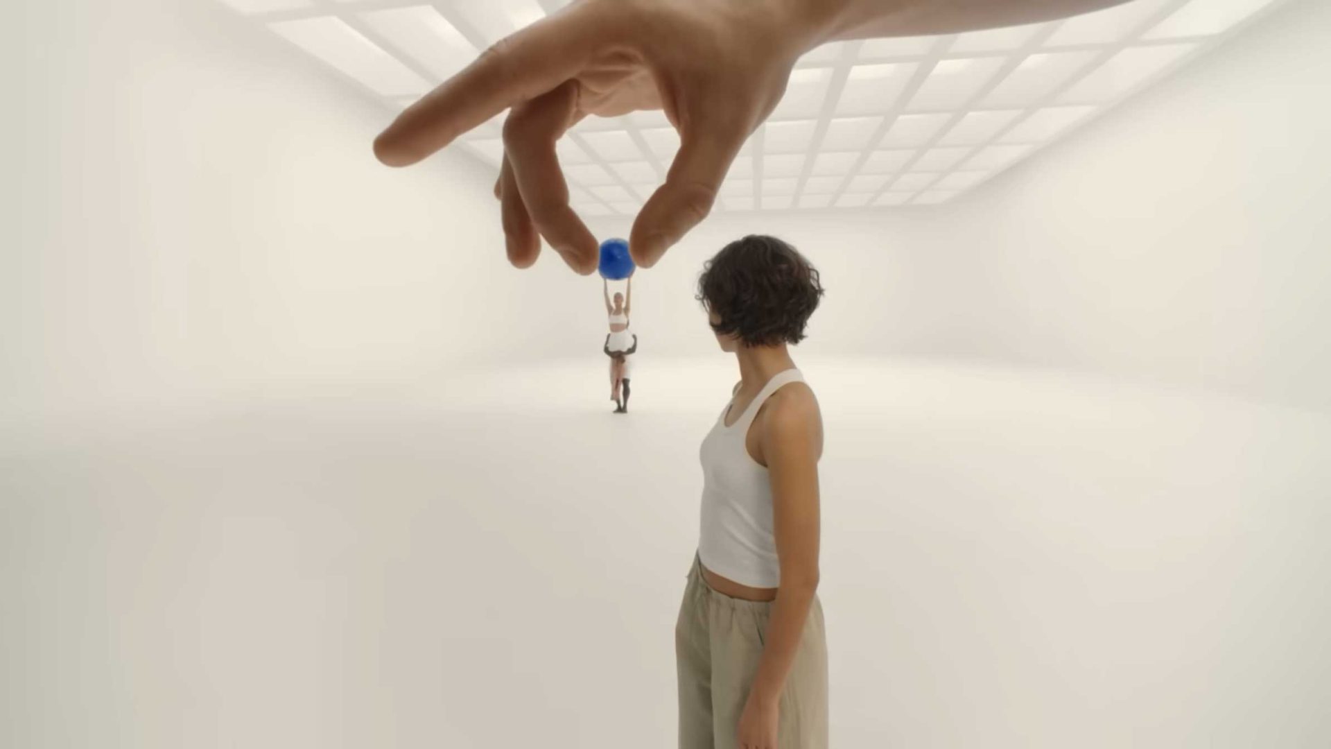 A person in the foreground gazes at a distant figure, who appears small and holds a blue ball. A large hand pinches the distant person amid the white, spacious room, evoking the surreal style of Spike Jonze’s Shape of Dreams. Frame from Stash Magazine article.