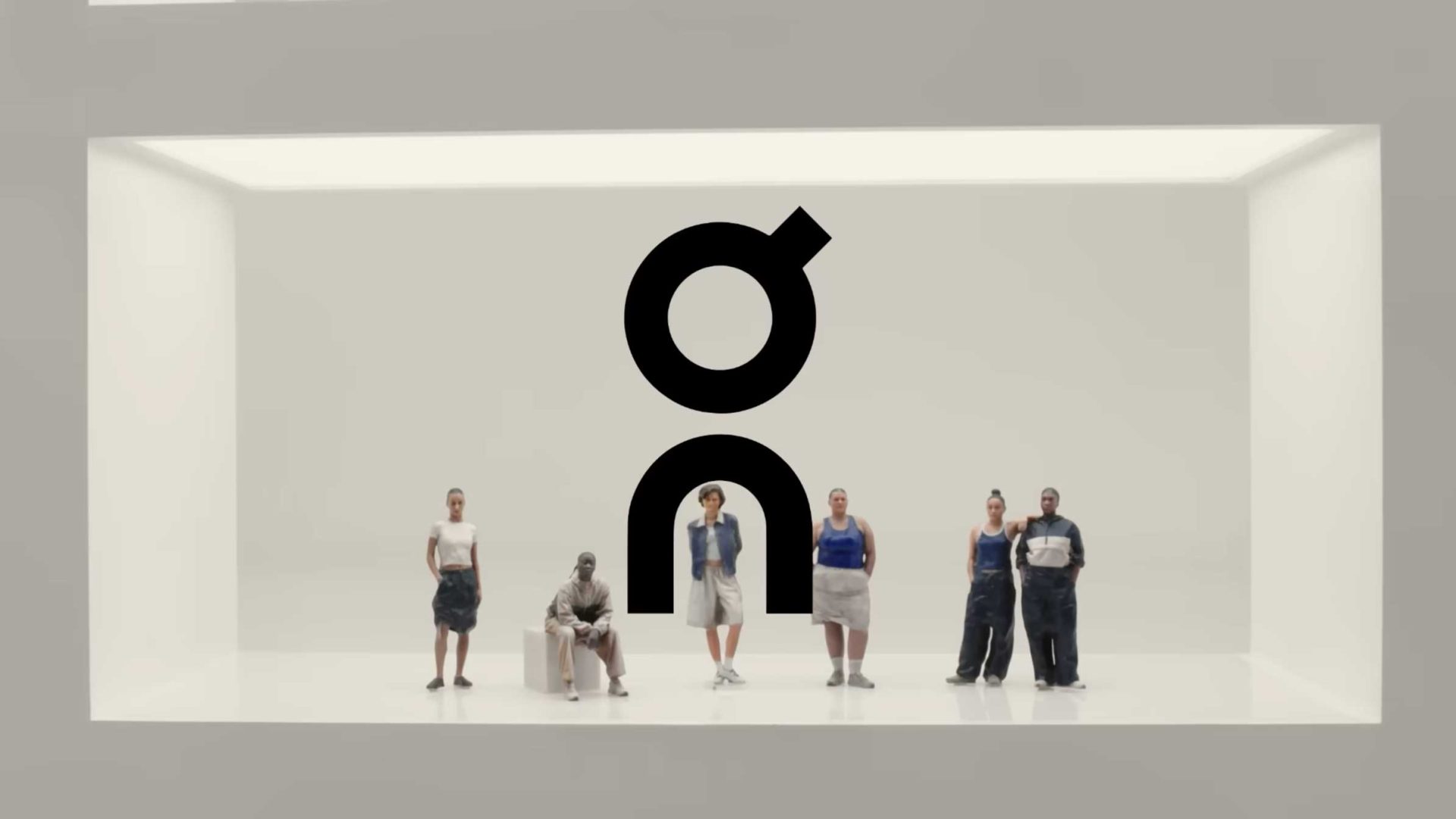 Six people, dressed in casual, sporty clothing, gather in a minimalist white room beneath a large black "On" logo. The scene evokes the Shape of Dreams, blending modern style with quiet energy. Frame from Stash Magazine article.