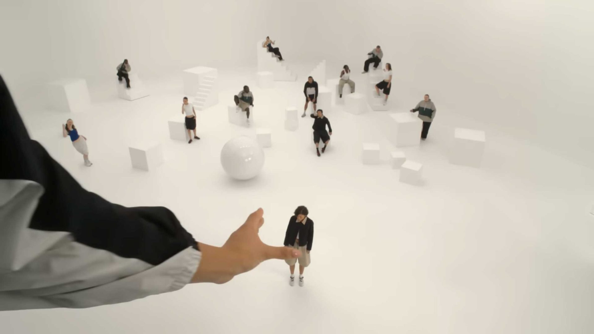 In a white, minimalistic room, several people sit or stand on geometric shapes. A large hand extends toward a central figure, creating a surreal effect—like a scene from the On Shape of Dreams ad featuring Zendaya, directed by Spike Jonze. Frame from Stash Magazine article.