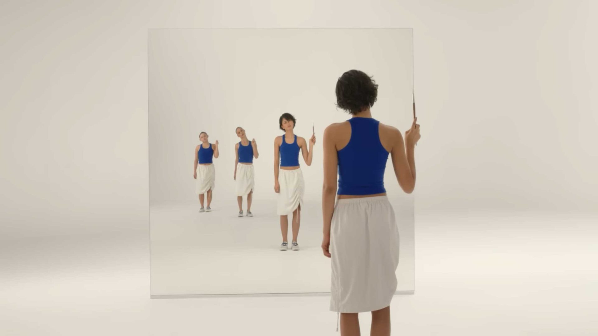 A person in a blue tank top and white skirt stands facing a large mirror, holding up one finger—like a scene from Spike Jonze's Shape of Dreams, the reflection shows multiple smaller versions disappearing into the distance. Frame from Stash Magazine article.