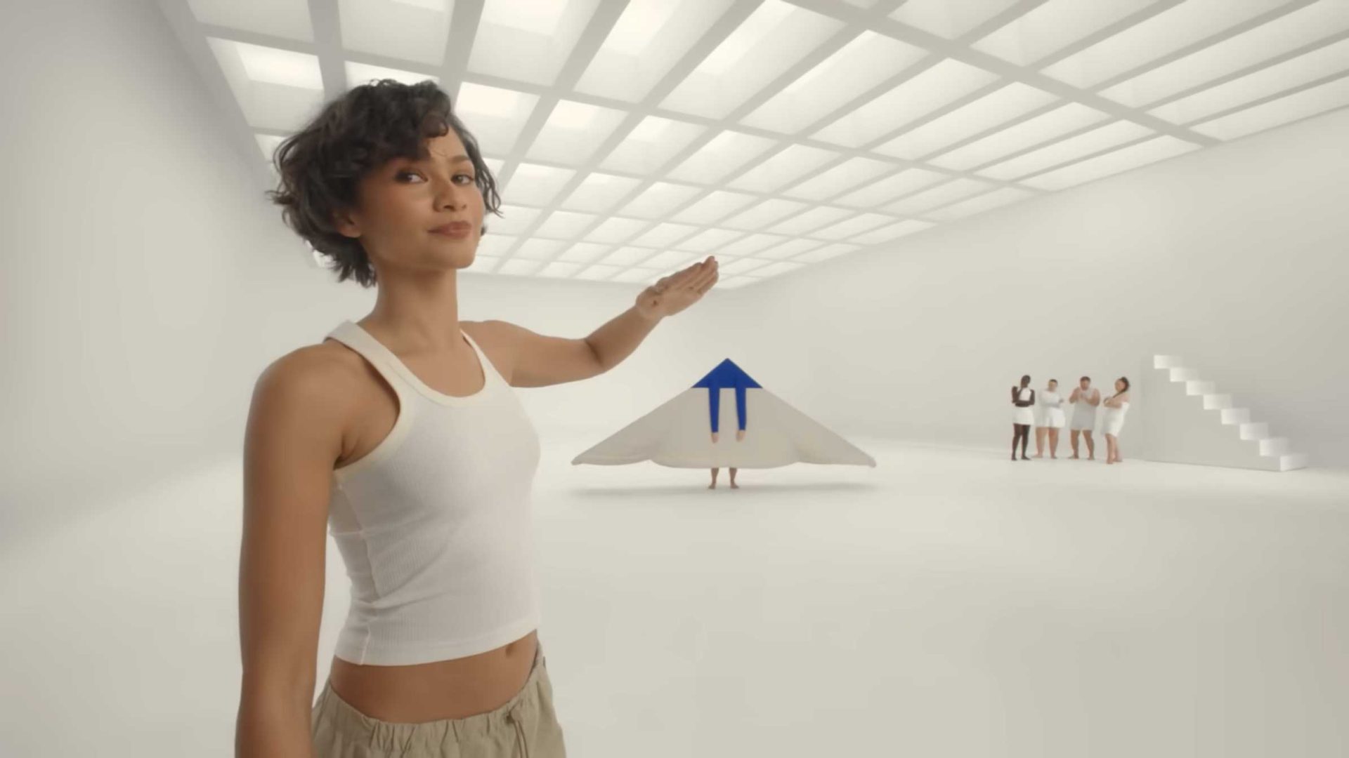 A woman in a white tank top gestures toward a person dressed as a white paper airplane with a blue arrow—an imaginative scene reminiscent of Spike Jonze. Four people stand in the background, all in a bright, minimalist white room with stairs on the right. Frame from Stash Magazine article.