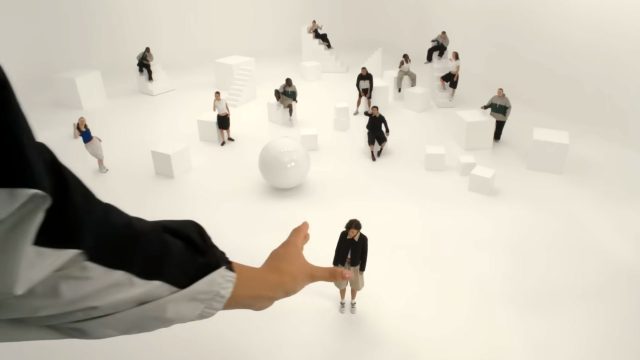 A large hand reaches toward a person standing alone in a white room filled with scattered cubes and spheres, evoking the surreal mood of Spike Jonze’s Shape of Dreams, while others sit or stand on white geometric shapes in the background. Frame from Stash Magazine article.