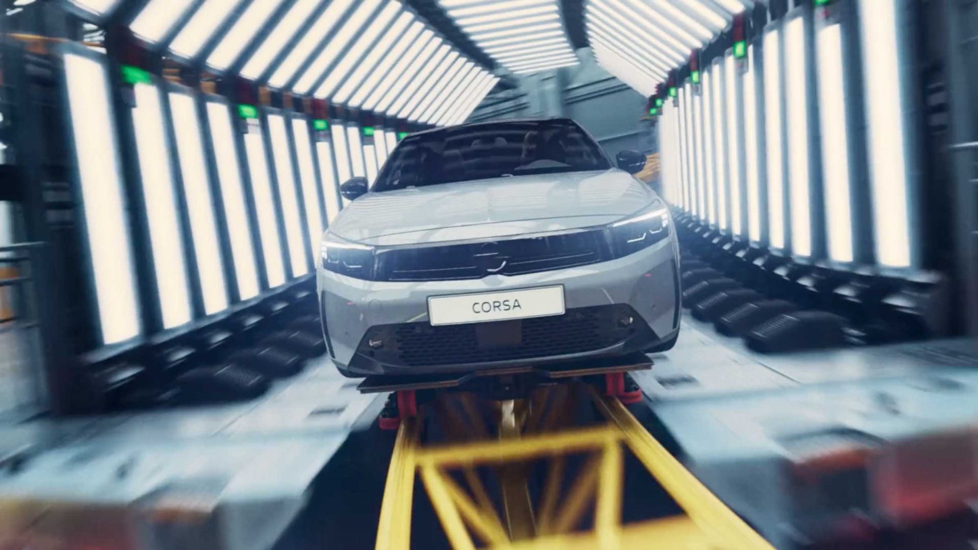 A silver Opel Corsa Coaster car is shown on a production line, surrounded by bright overhead and side lights in a modern automotive factory, highlighting the work of designer Mark Jenkinson. Frame from Stash Magazine article.