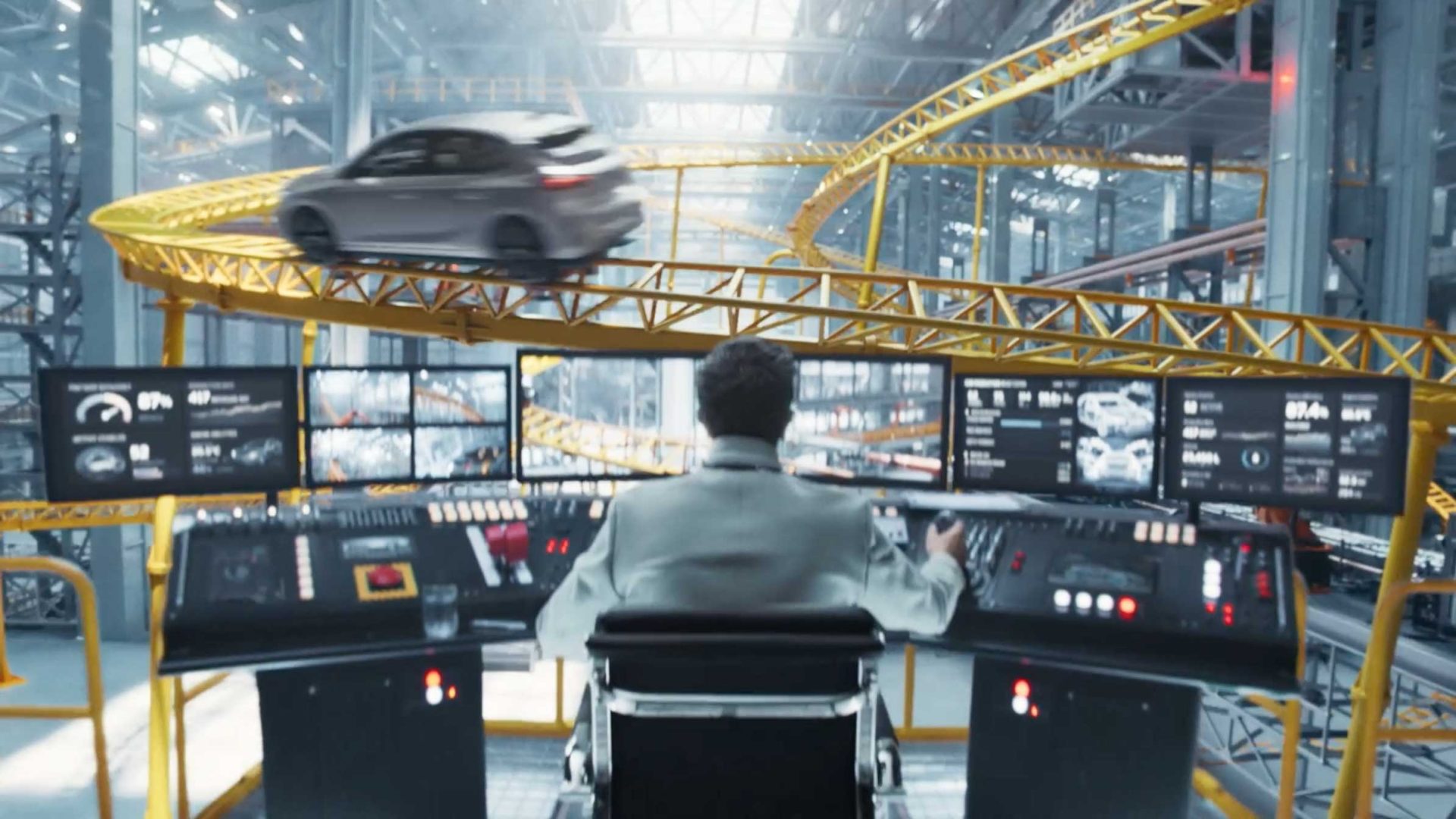 A person sits at a control desk with multiple monitors in a high-tech facility, watching the Opel Corsa Coaster speed along a yellow elevated track that winds through a large, industrial indoor space. Frame from Stash Magazine article.