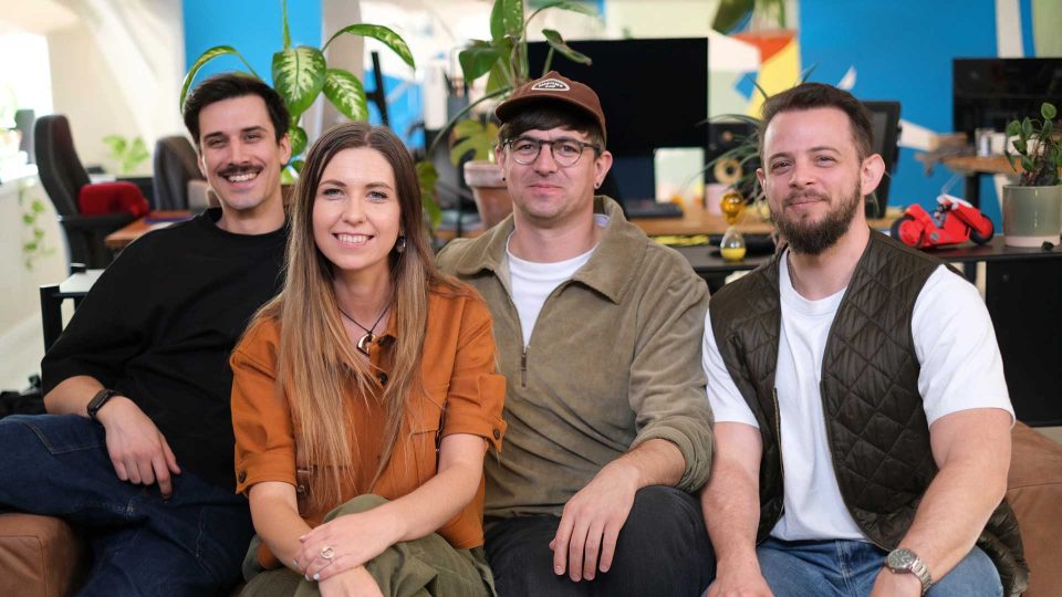 Four people sitting closely together and smiling at the camera in a bright, colorful office with plants and desks, celebrating as Panic Animation Studio acquires Kingdom of Something. Frame from Stash Magazine article.