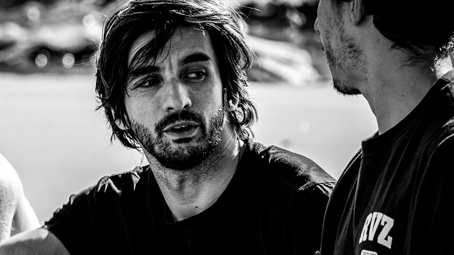Black and white photo of two men talking outdoors. One man with tousled hair and a beard looks intently at the other, who is partly turned away—a candid moment as Papaya-Films-sign-director--Favio-Vinson-to-roster. The background is blurred. Frame from Stash Magazine article.