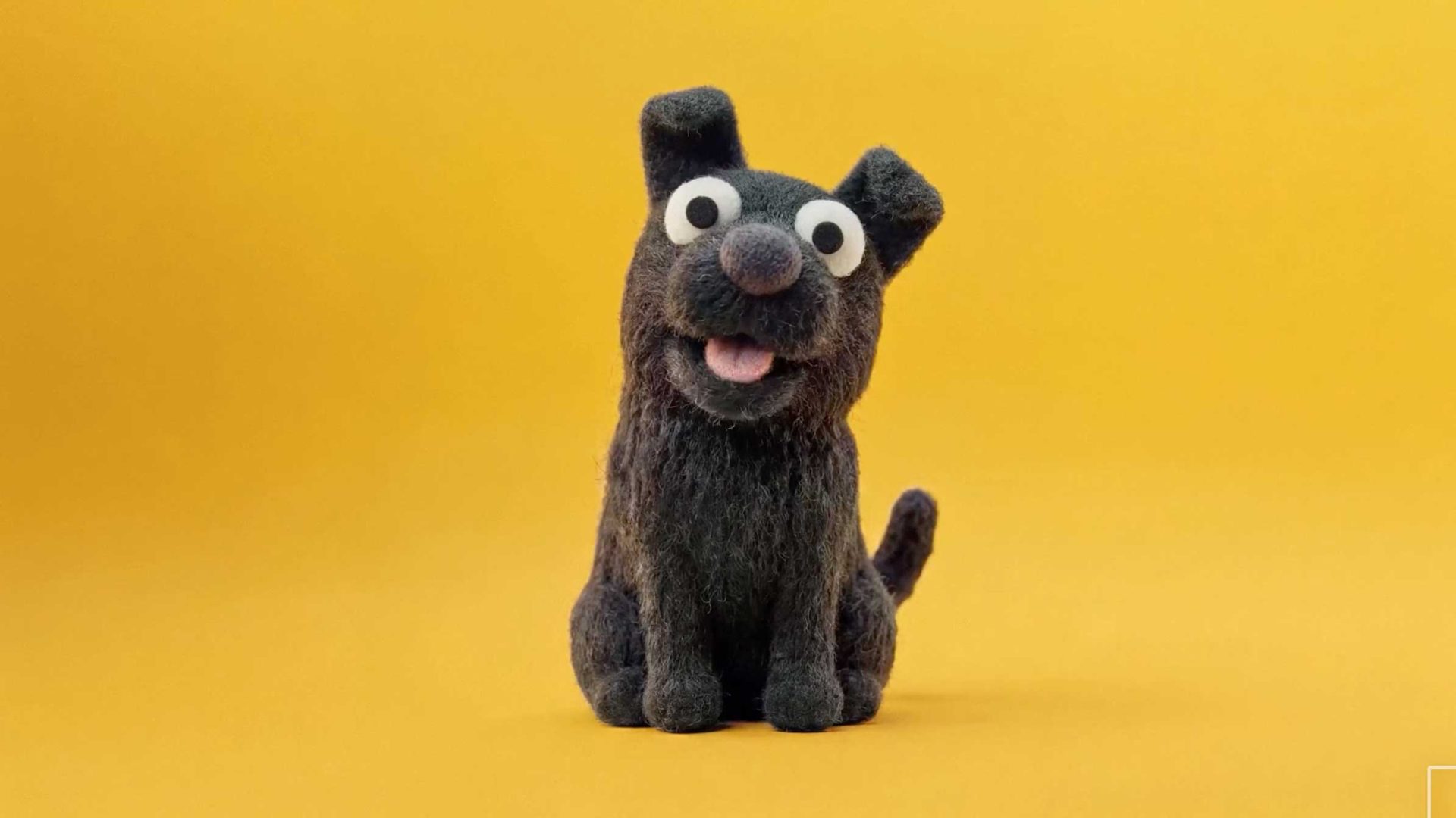 A felted black puppy figurine with large googly eyes sitting against a yellow background, captured from a pet POV. Frame from Stash Magazine article.