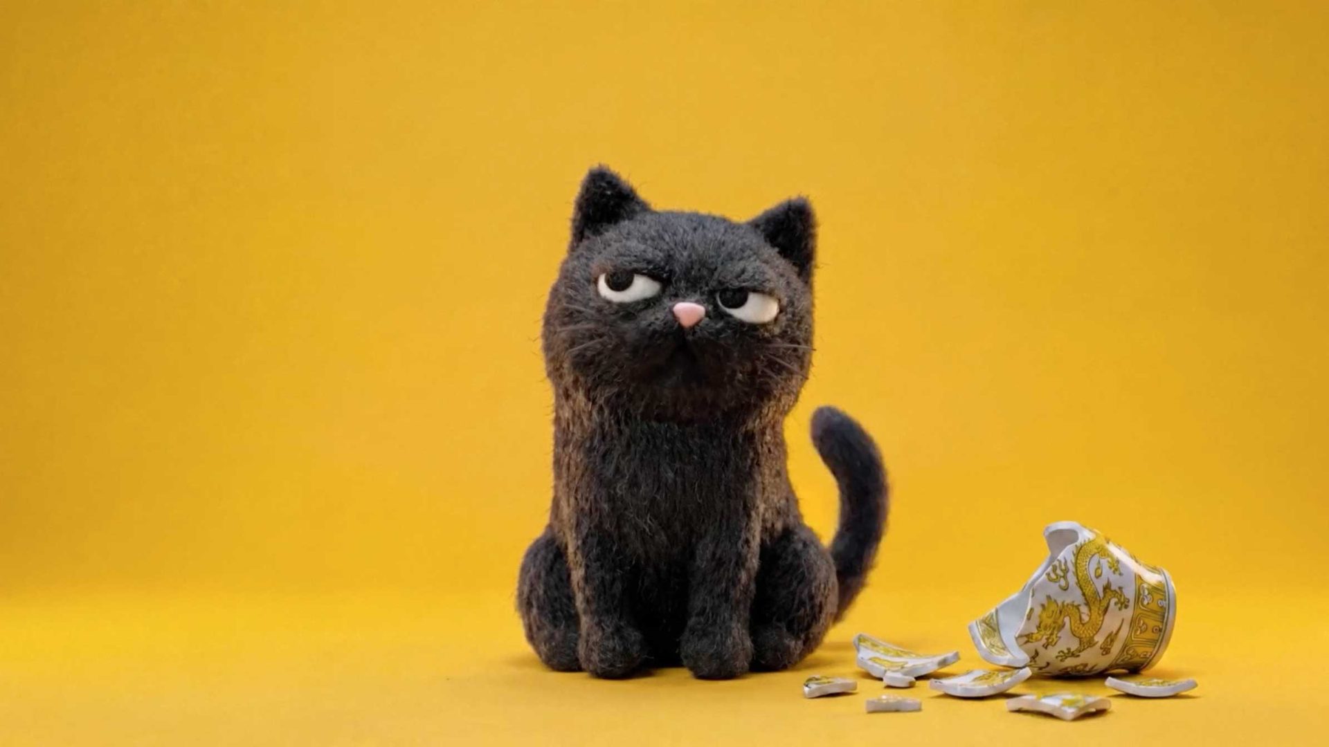 A needle-felted black cat from the Petbarn Petey and Barnadette brand films sits with a grumpy expression next to a broken ceramic cup. Frame from Stash Magazine article.