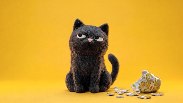 A grumpy-looking felted black cat sits next to a shattered ceramic cup on a yellow background, reminiscent of the Petey and Barnadette brand films. Frame from Stash Magazine article.