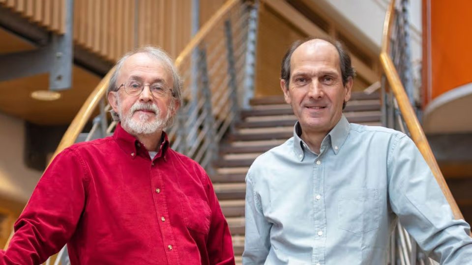 Peter Lord and David Sproxton, co-founders of Aardman Animations, stand indoors by a wooden staircase. One wears a red shirt and glasses with a white beard; the other is in light blue. Both look at the camera, with the railing behind them. Frame from Stash Magazine article.