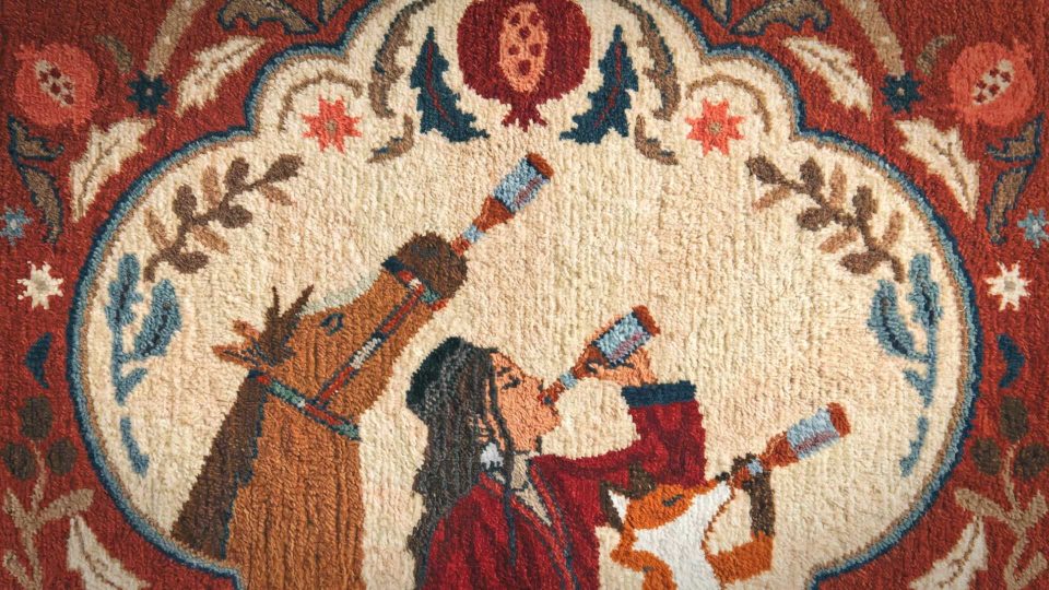 A woven tapestry shows a person and two horses, all looking through telescopes. The person and horses are side by side, with ornate, colorful designs framing them in shades of red, gold, and blue—a striking visual that echoes how Petrick & Director Natalia Bazina roll out the carpet for WOSTOK. Frame from Stash Magazine article.