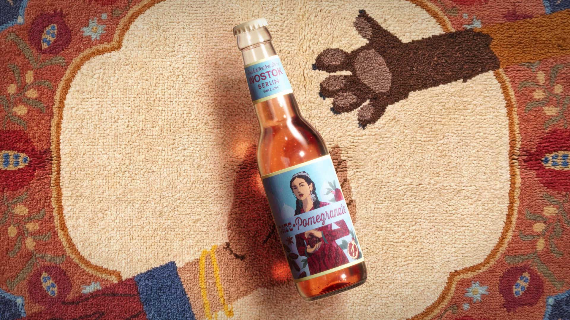 A bottle of WOSTOK pomegranate soda with a vintage-style label lies on a colorful, patterned rug featuring a cartoon bear’s face and paw. Frame from Stash Magazine article.