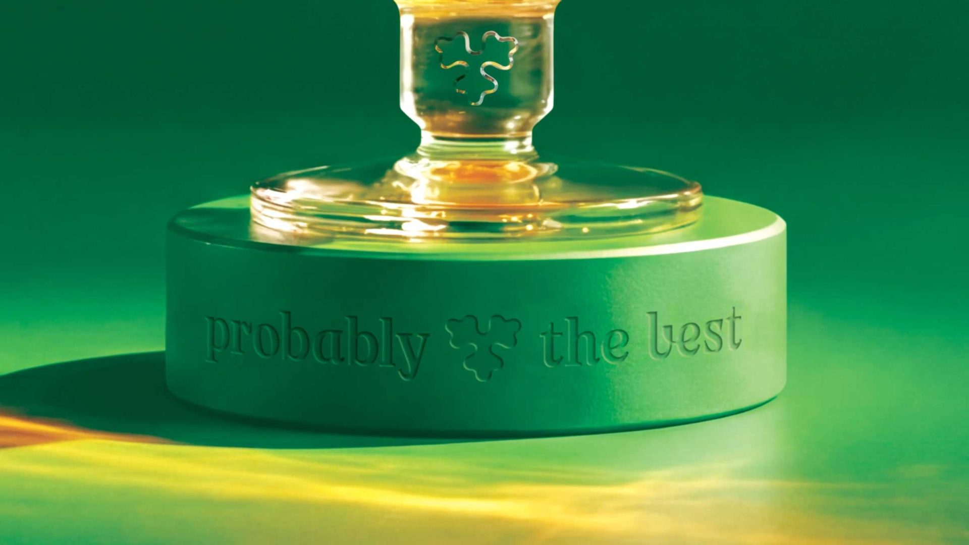 A close-up of a green pedestal with a glass object on top. The pedestal, engraved in Probably Serif font by Taxi Studio with the words “probably the best,” features a clover-shaped logo against a green-to-yellow gradient background. Frame from Stash Magazine article.