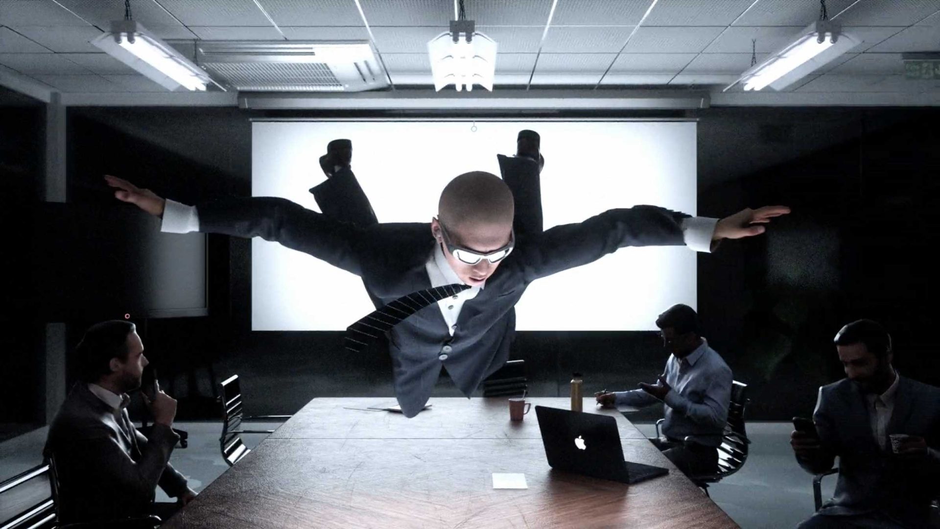 A man in a suit dives superhero-style over a conference table while three others sit around, focused on their laptops and phones in a modern office meeting room—a scene reminiscent of Quentin Deronzier’s dynamic visuals, capturing The Art of Losing It amid corporate chaos. Frame from Stash Magazine article.