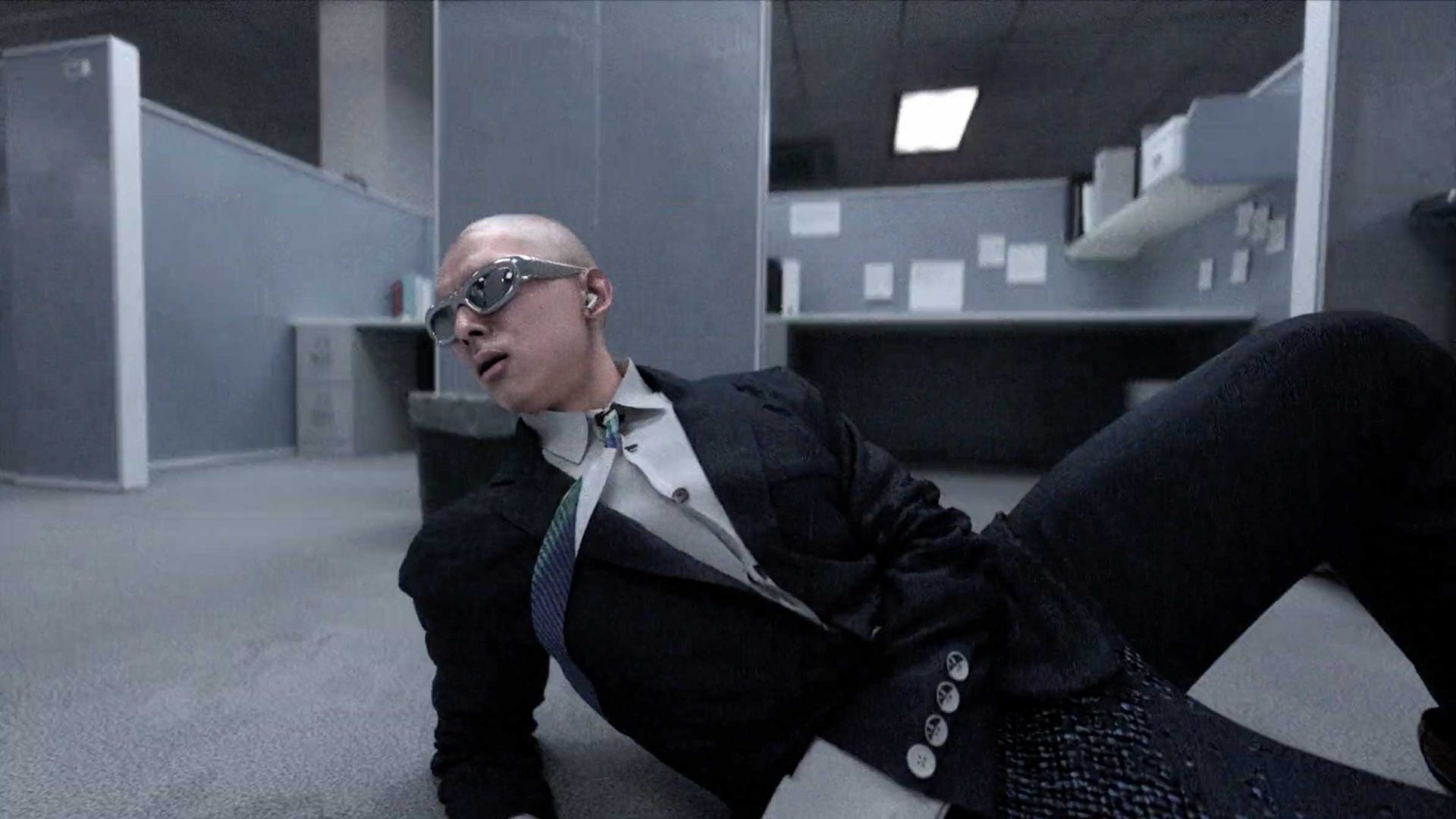 A person in a suit, wearing sunglasses and an earpiece, is lying on the floor of a modern office with cubicles, looking off to the side with a surprised expression—capturing a scene that echoes Quentin Deronzier’s surreal aesthetics and themes from The Art of Losing It. Frame from Stash Magazine article.