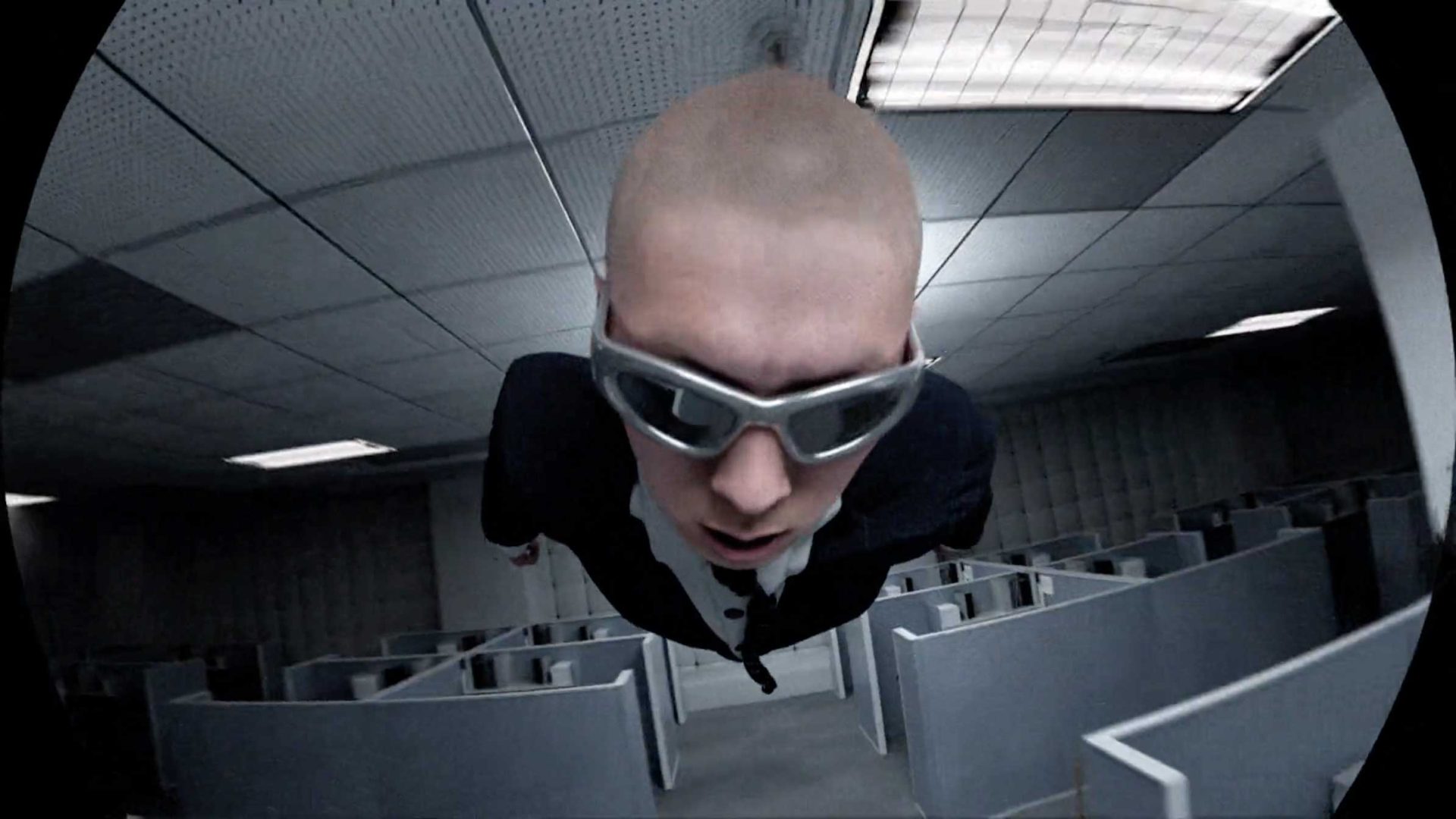A person in a suit and sunglasses is floating horizontally above cubicles in an office, viewed from a fisheye perspective with a distorted ceiling and fluorescent lights—a surreal scene reminiscent of Quentin Deronzier’s visual style, perfectly capturing The Art of Losing It. Frame from Stash Magazine article.