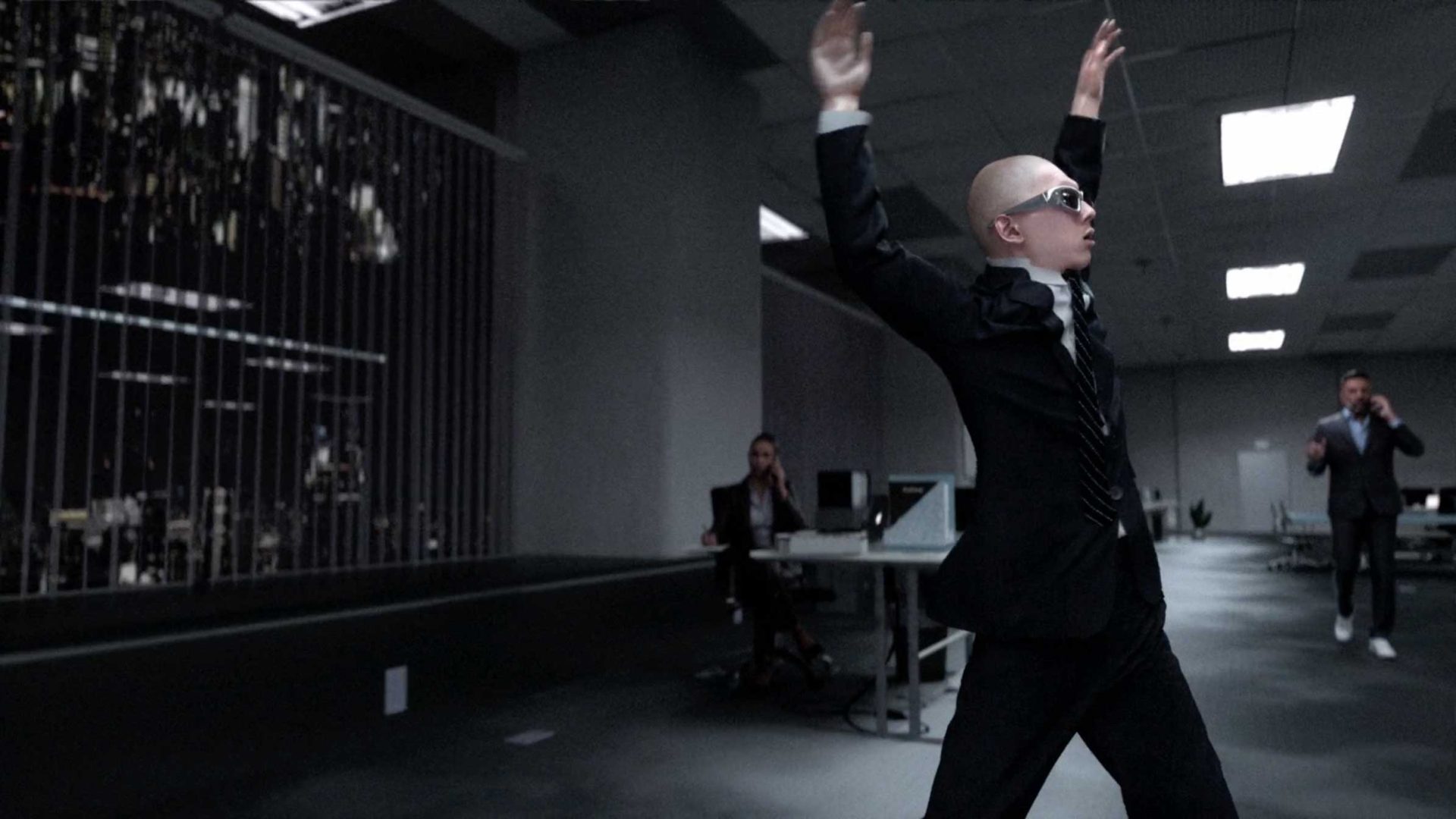 A person in a suit and sunglasses raises their arms dramatically in an office setting, channeling the bold energy of The Art of Losing It, while others at desks watch. The modern office boasts a striking cityscape through large windows, adding a cinematic flair reminiscent of Quentin Deronzier’s visual style. Frame from Stash Magazine article.