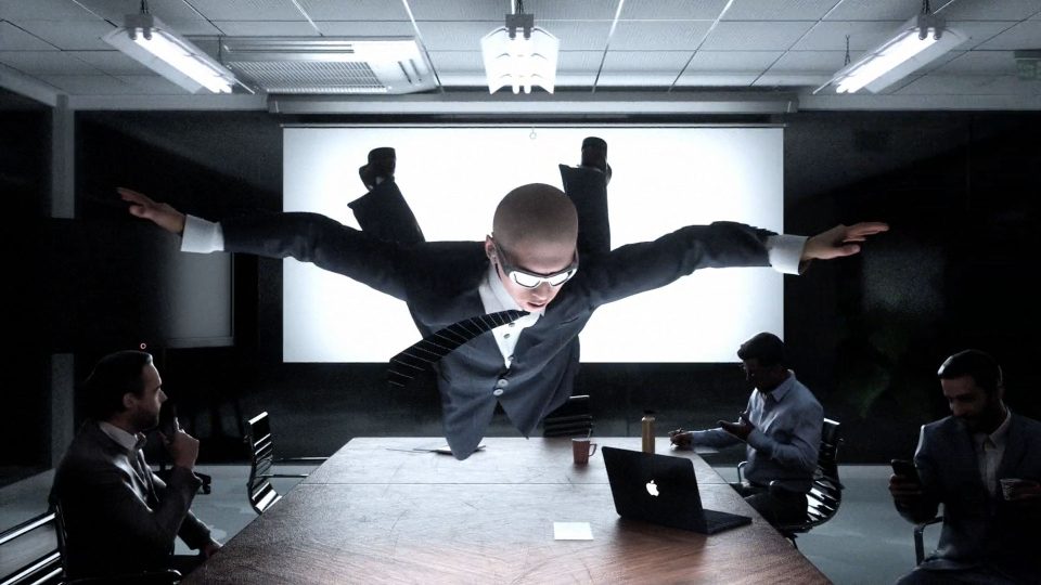 A man in a suit and glasses is suspended horizontally over a conference table, arms outstretched, while three seated men watch or work on laptops in a modern meeting room—evoking the surreal visual style of Quentin Deronzier’s short film The Art of Losing It. Frame from Stash Magazine article.