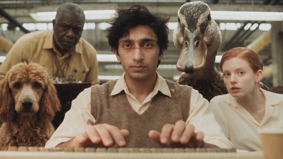 A man types on a keyboard in a scene from the short film Query, flanked by a poodle, an older man, a large duck, and a woman. Frame from Stash Magazine article.
