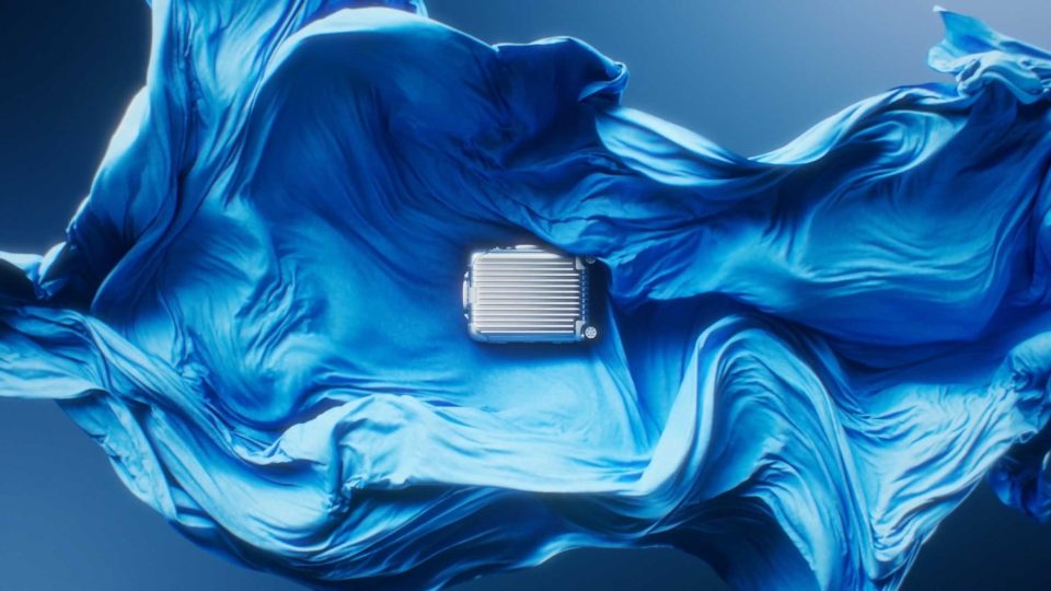 A small, metallic silver RIMOWA suitcase is centered on flowing, textured blue fabric moving like a wave, against a softly lit blue background in this "Precision in Motion" spec film by Solve Studios. Frame from Stash Magazine article.