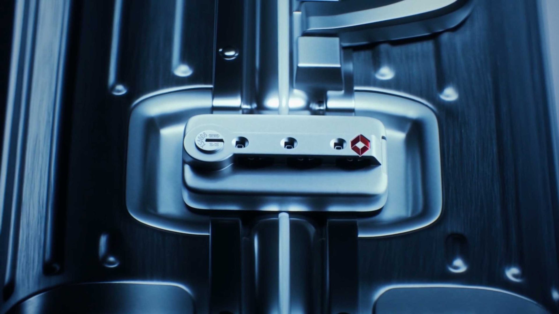 Close-up of a silver TSA-approved combination lock on a RIMOWA hard-shell suitcase, showcasing brushed metal texture and the red diamond-shaped Travel Sentry logo—Precision in Motion for discerning travelers. Frame from Stash Magazine article.