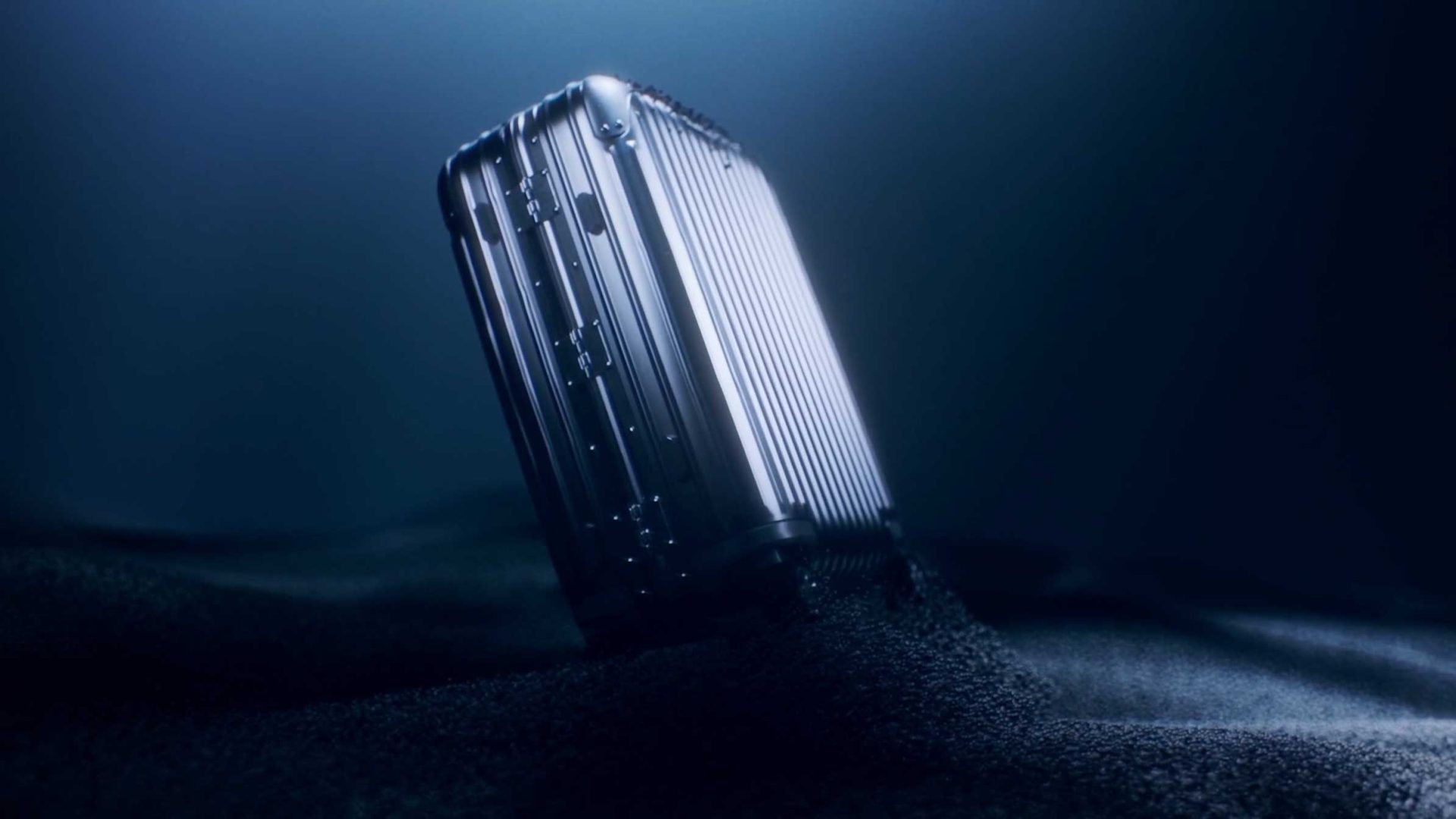 A metallic, ridged RIMOWA suitcase stands upright, partially buried in dark sand and illuminated by a dramatic blue light—"Precision in Motion" spec film by Solve Studios evokes a futuristic and mysterious atmosphere. Frame from Stash Magazine article.