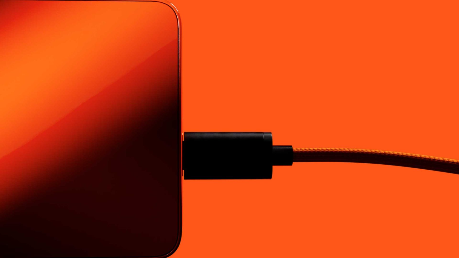 A close-up of a black charging cable plugged into the side of a Rabbit r1 device, set against a vibrant orange background—ideal for a product film by Styleframe GmbH. Frame from Stash Magazine article.