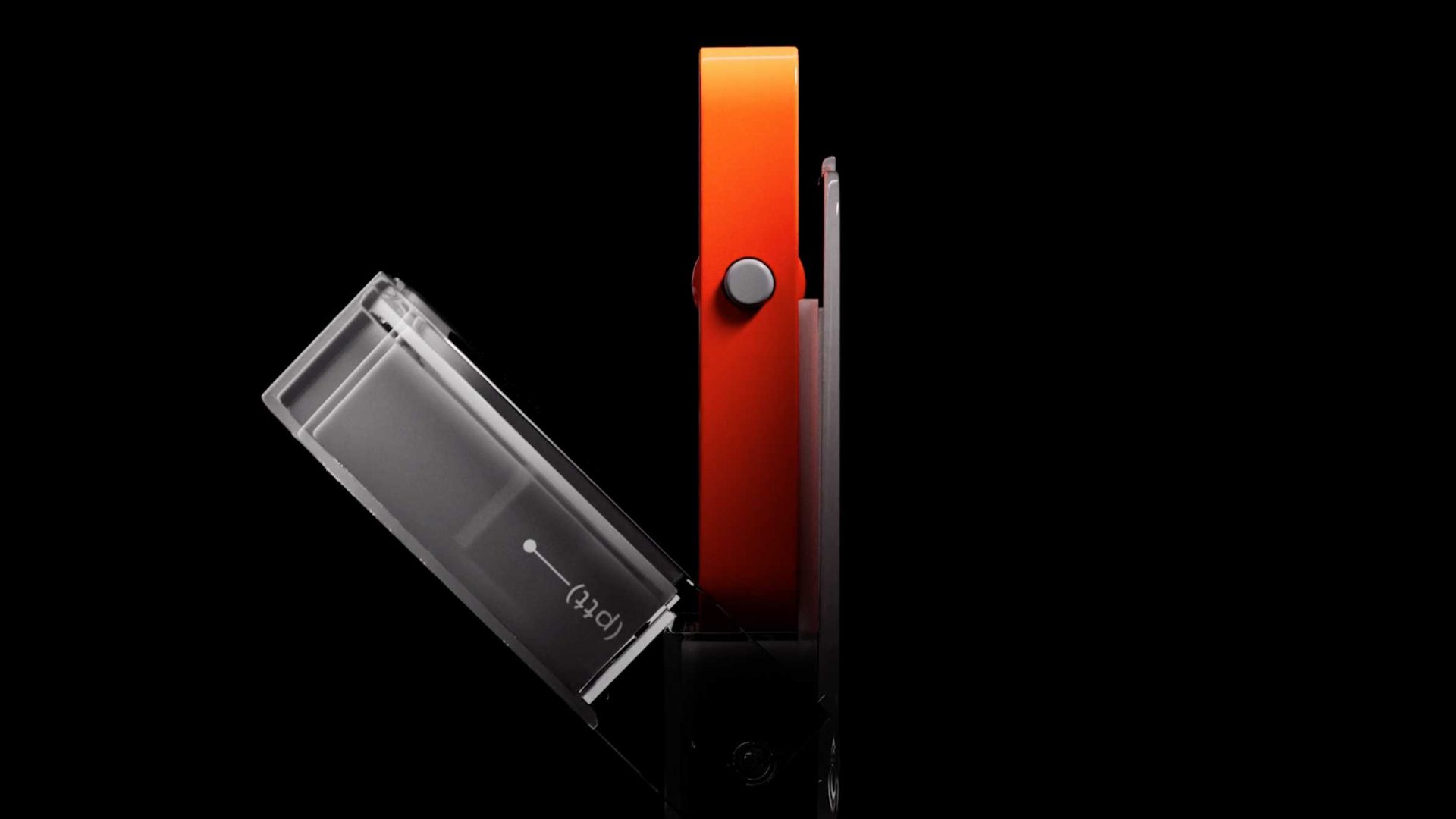 Side view of two objects against a black background: a transparent cassette tape tilted open next to an orange Rabbit r1 product film device by Styleframe GmbH, both aligned vertically with a round black button. Frame from Stash Magazine article.