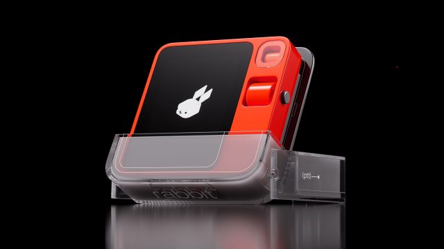 A compact, orange and black electronic device with a white rabbit logo on its screen sits in a transparent charging dock against a black background, featured in the Rabbit r1 product film by Styleframe GmbH. Frame from Stash Magazine article.