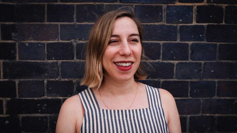 A woman wearing a sleeveless, striped top stands in front of a dark brick wall, smiling and looking slightly to the side—exuding the poised confidence of Rebecca_Scheinberg, executive producer at ETC. Frame from Stash Magazine article.