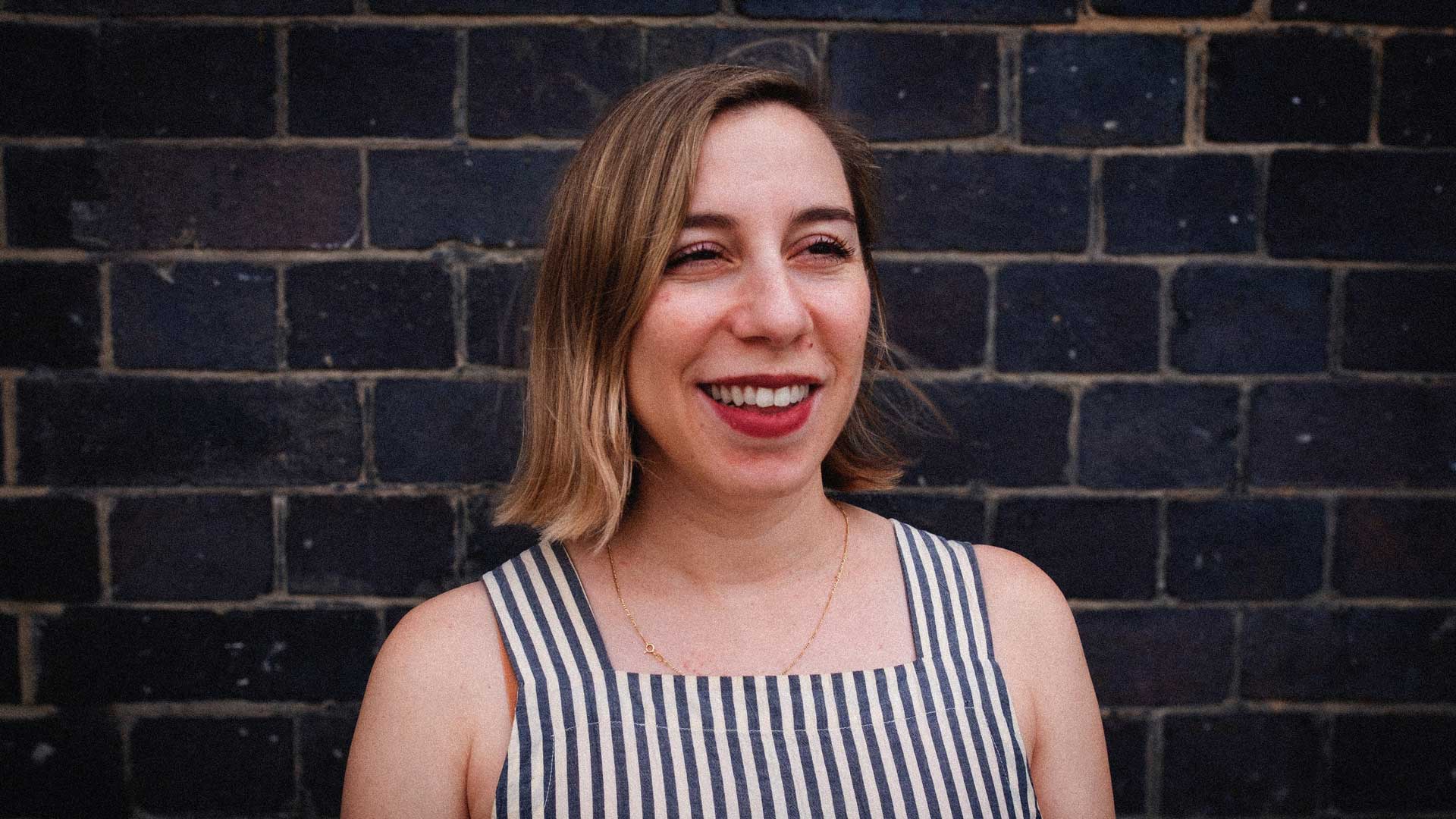 A woman wearing a sleeveless, striped top stands in front of a dark brick wall, smiling and looking slightly to the side—exuding the poised confidence of Rebecca_Scheinberg, executive producer at ETC. Frame from Stash Magazine article.