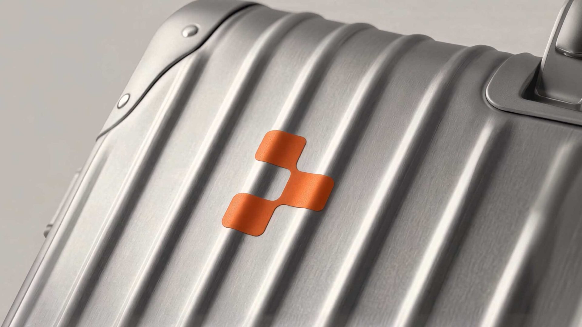 A close-up of a silver hard-shell suitcase with a matte finish, featuring an orange geometric logo inspired by Replit in the center. The suitcase has ridged vertical lines and metallic hardware. Frame from Stash Magazine article.
