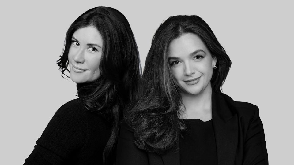 Two women with long, dark hair pose back-to-back, both smiling slightly. They are wearing dark clothing and are set against a plain, light grey background. The image is in black and white. Frame from Stash Magazine article.