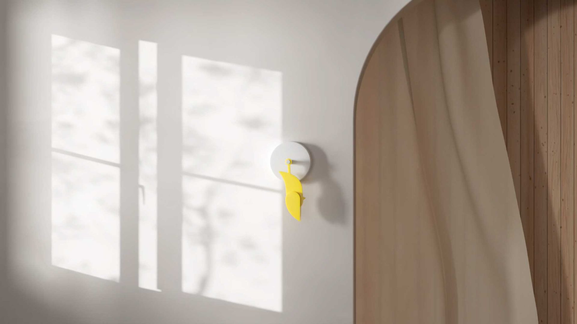 Minimalist interior with sunlight streaming through a window, casting shadows on a white wall. A modern Ribbit wall clock with a bright yellow pendulum is mounted near a wooden arched doorway, evoking Copenhagen-inspired design. Frame from Stash Magazine article.