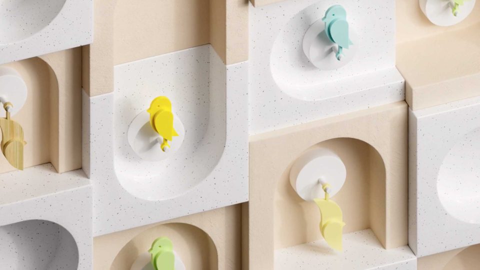 Colorful bird-shaped hooks in yellow, green, and blue are mounted on a patterned wall with curved and arched niches in beige and white tones—a minimalist, geometric scene inspired by Ribbit Launches in Copenhagen With Birdie Brand Film. Frame from Stash Magazine article.