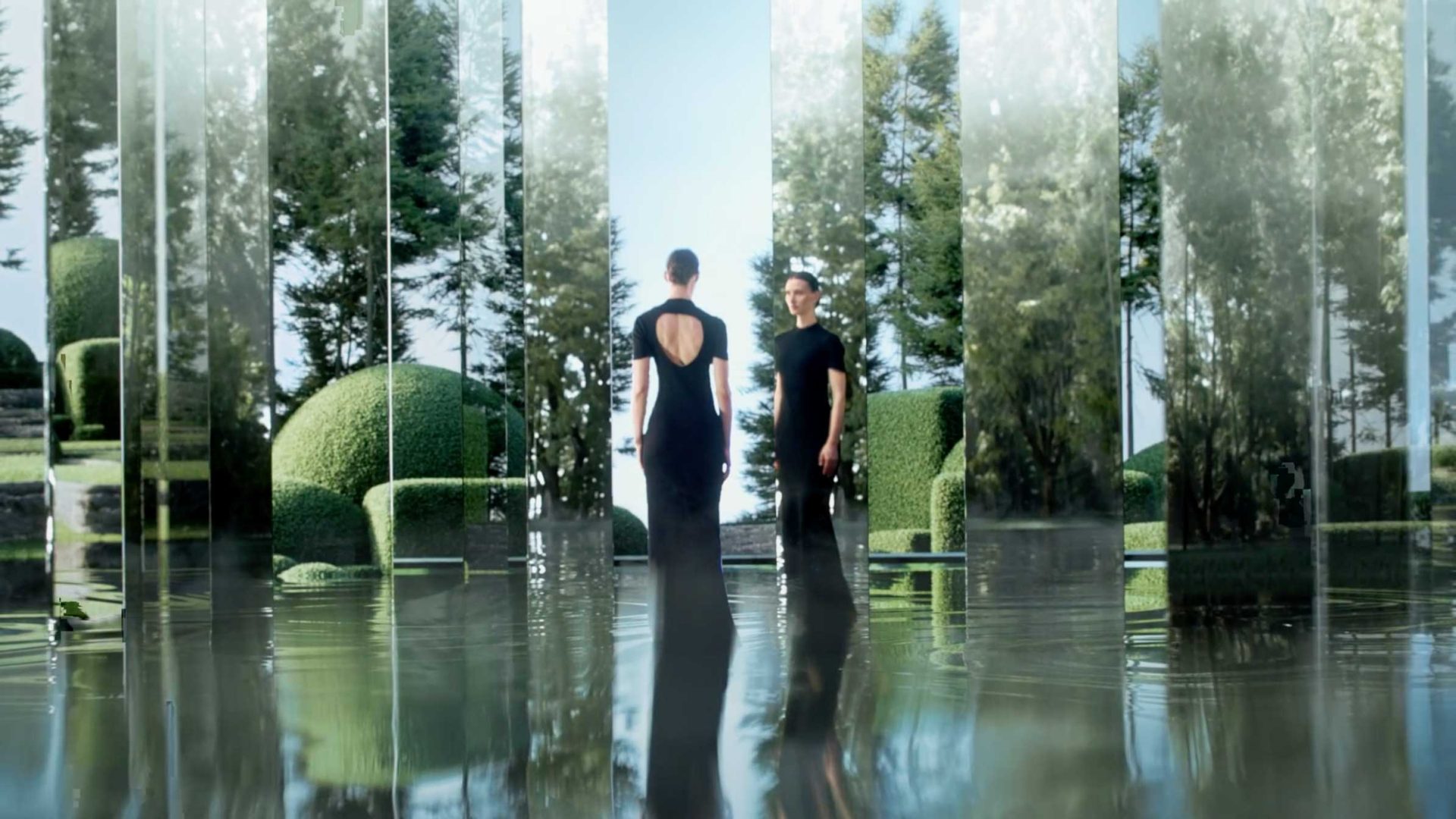 Two people in black outfits stand among tall mirrored panels in a modern garden, their reflections creating a surreal, symmetrical effect reminiscent of Roger Dubuis elegance on a shiny surface amid trimmed bushes and trees. Frame from Stash Magazine article.