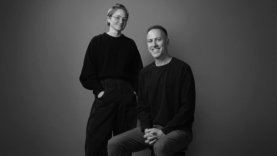 A woman stands with hands in pockets next to a seated man. Both wear dark clothing and smile at the camera against a plain, neutral background, celebrating Versus bolstering leadership ranks with ECD Rosie Garschina & EP Kevin Anderson. The image is in black and white. Frame from Stash Magazine article.