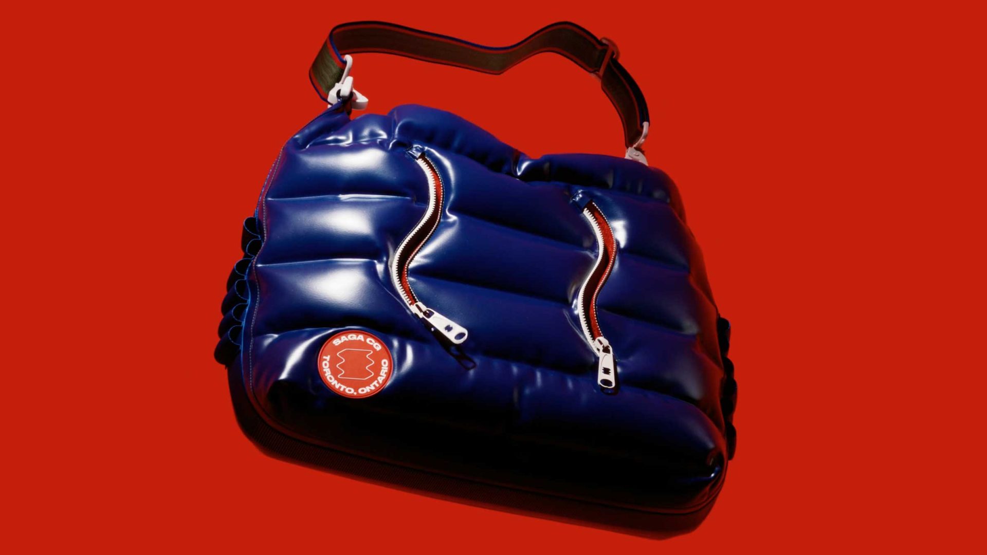 A shiny, padded dark blue shoulder bag with two silver zippers and an adjustable strap, featuring a red and white circular patch inspired by the SAGA studio launch film, set against a solid red background. Frame from Stash Magazine article.