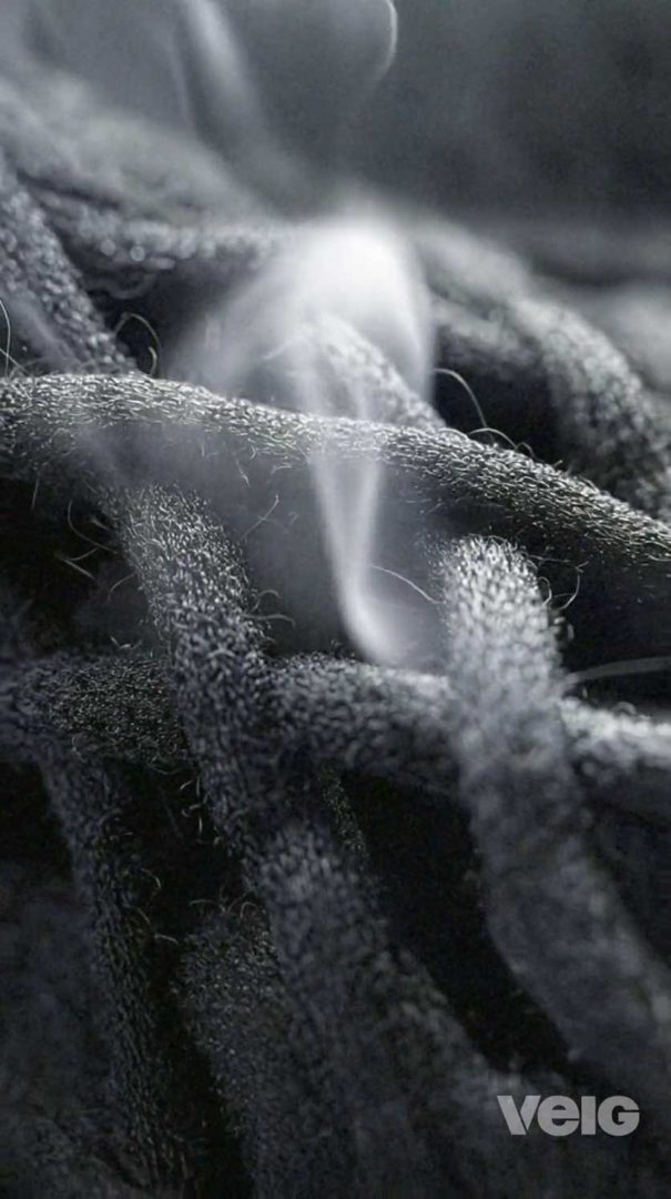 Close-up of textured, dark fabric from the TENAYA PACK 45L with wisps of steam or smoke rising from the surface. The logo VELG appears in the lower right corner. Frame from Stash Magazine article.