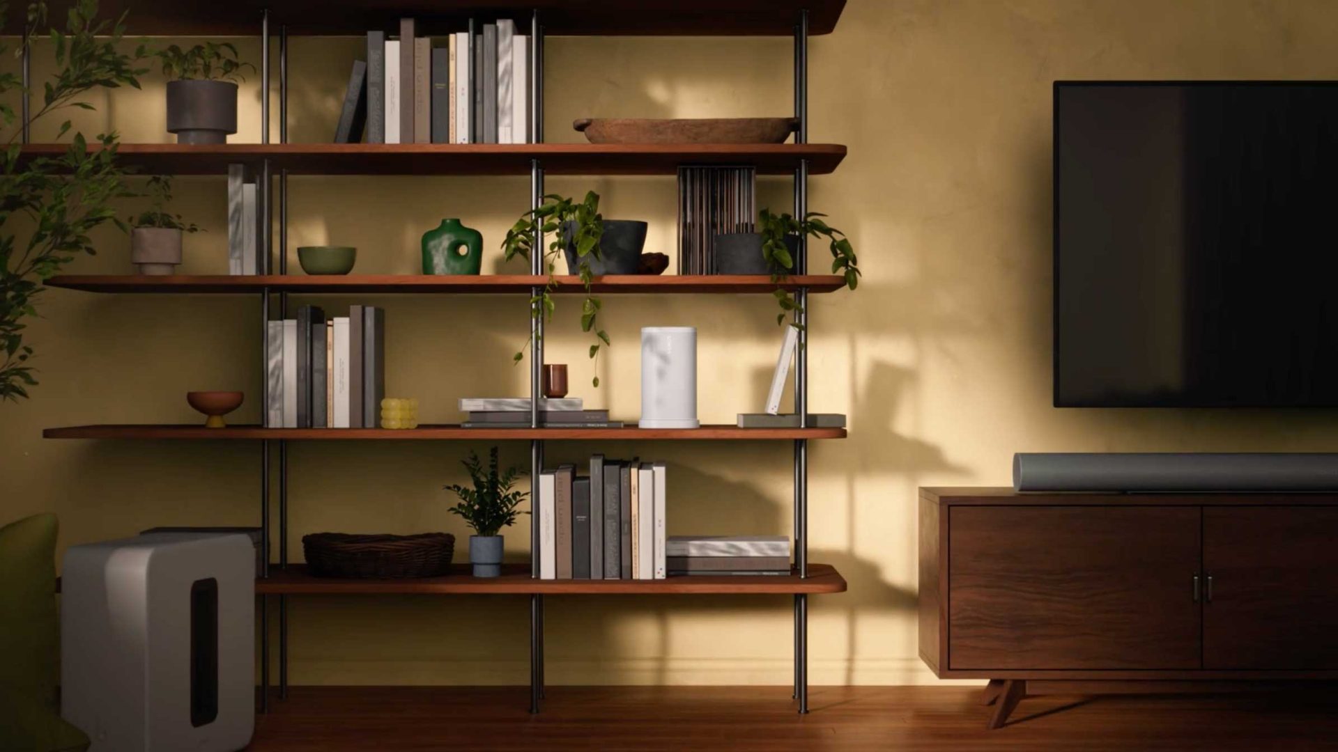 A modern living room with wooden shelves holding books, plants, and decorative objects. A Sonos Play speaker sits beside the flat-screen TV mounted on the wall above a wooden cabinet. Warm lighting casts soft shadows on the yellow wall. Frame from Stash Magazine article.