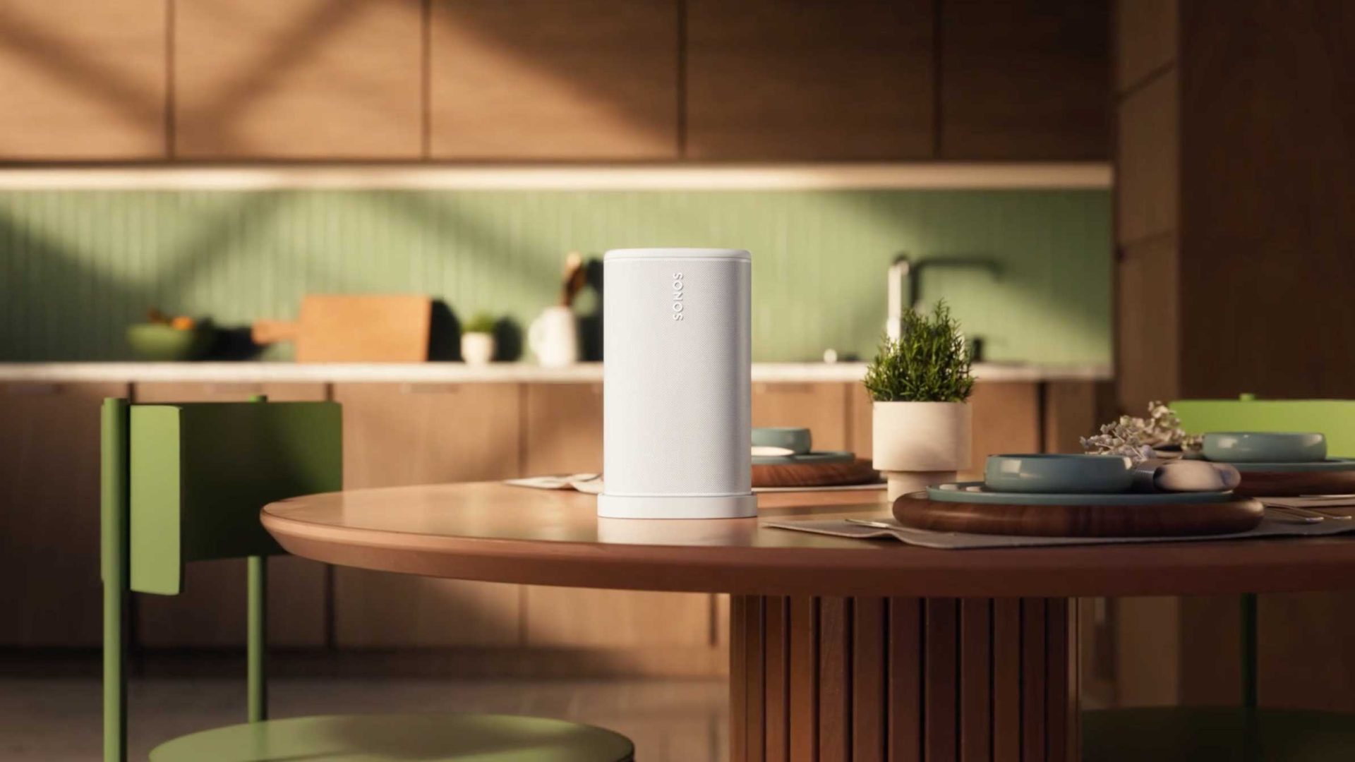 A white cylindrical speaker, inspired by the Sonos “Play” brand film by Frame48, sits on a round wooden dining table set with plates, cutlery, and a small plant in a modern kitchen with green chairs and wooden cabinets. Frame from Stash Magazine article.