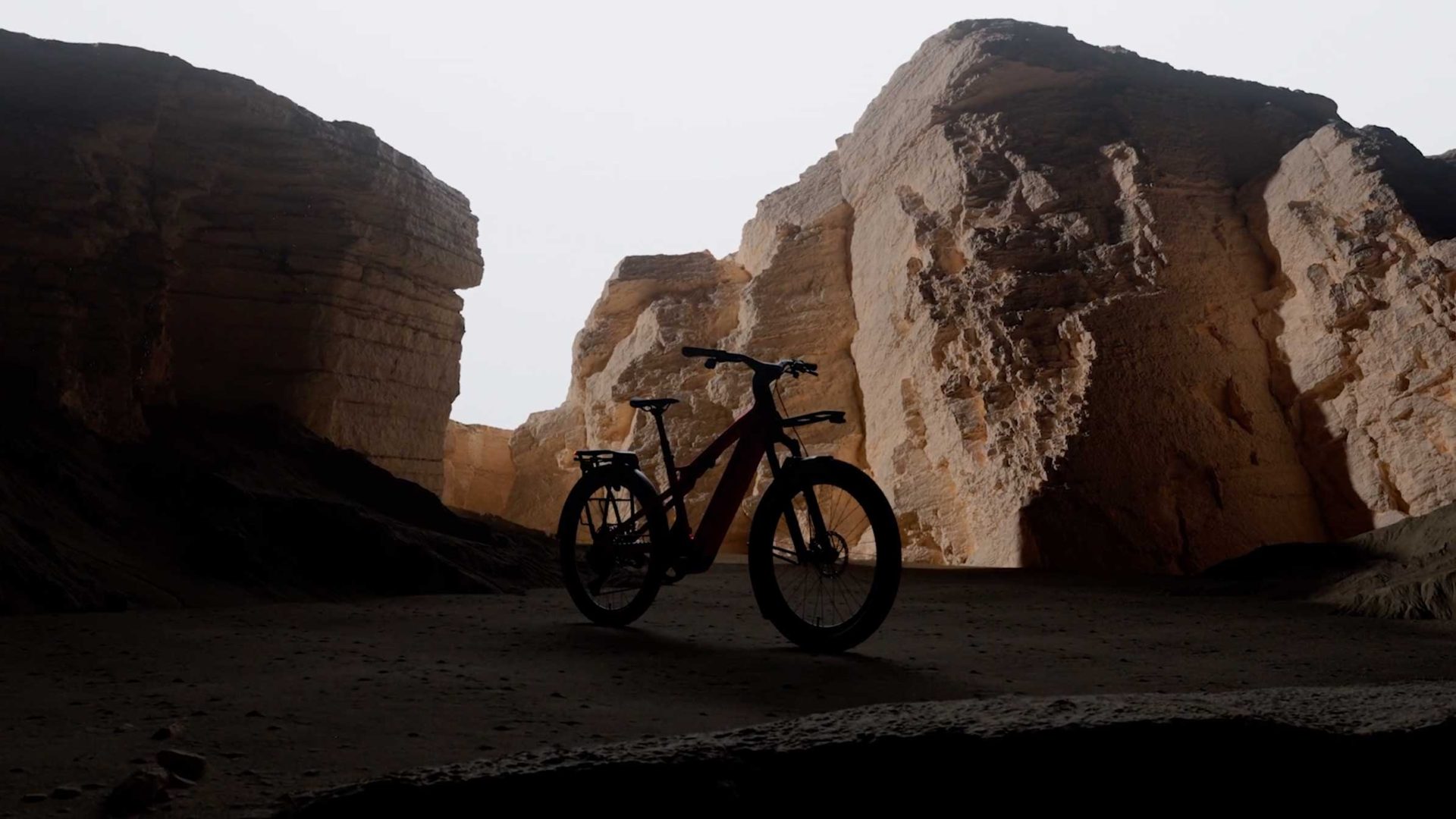 A Specialized Vado 3 launch by Inertia Studios features a bicycle silhouetted in a dark cave, framed by large, sunlit rock formations and cliffs in the background. Frame from Stash Magazine article.