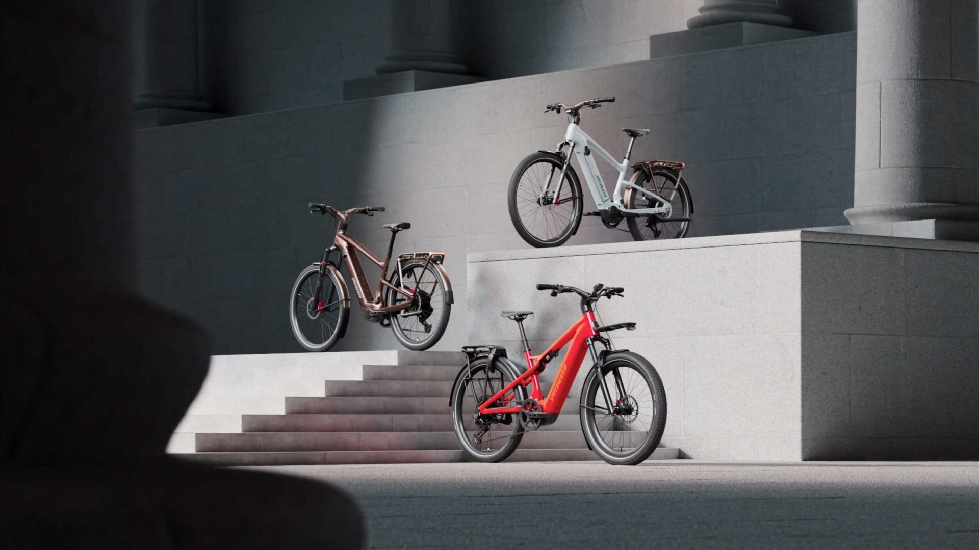 Three electric bicycles—including the Specialized Vado 3 in brown, white, and red—are displayed on different levels of wide stone steps against a grey building with columns, illuminated by angled sunlight. This dynamic arrangement captures a sense of inertia and excitement reminiscent of a product launch. Frame from Stash Magazine article.