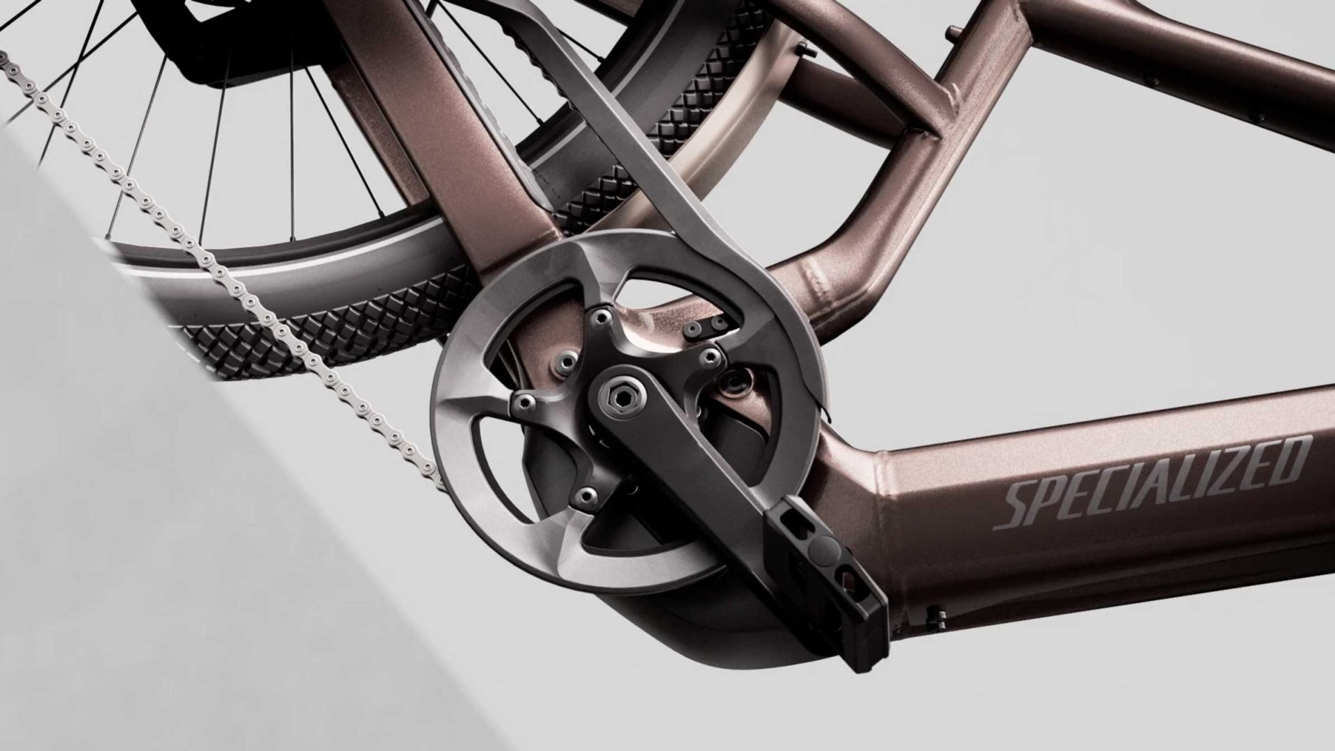 Close-up photo of a bronze-colored bicycle frame, focusing on the crankset, pedal, and chain, with the word SPECIALIZED visible on the frame. This image captures the precise engineering of the Specialized Vado 3 at launch, highlighting its refined design and sense of inertia against a light gray and white background. Frame from Stash Magazine article.